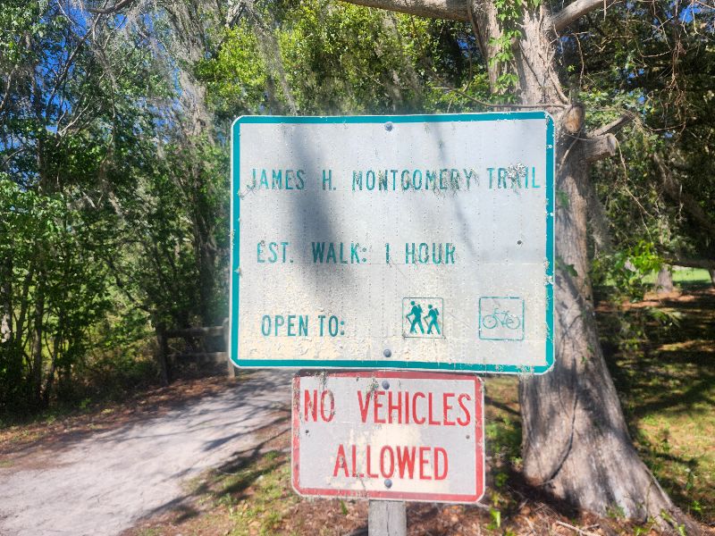 A sign for the James H. Montgomery Trail stands before a wooded path featuring trees covered in Spanish moss. Estimated walk time for the trail is 1 hour. Pedestrians and biks are allowed but no vehicles.